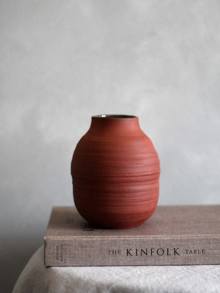 Handgemaakte bloemenvaas van rode klei, op een boek, op een tafekleed met een grijze achtergrond