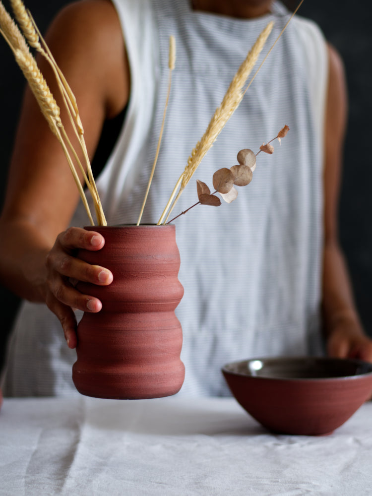 Rode bloemenvaas met golven, vastgehouden door een hand, met een rode kom vaag op de achtergrond. 