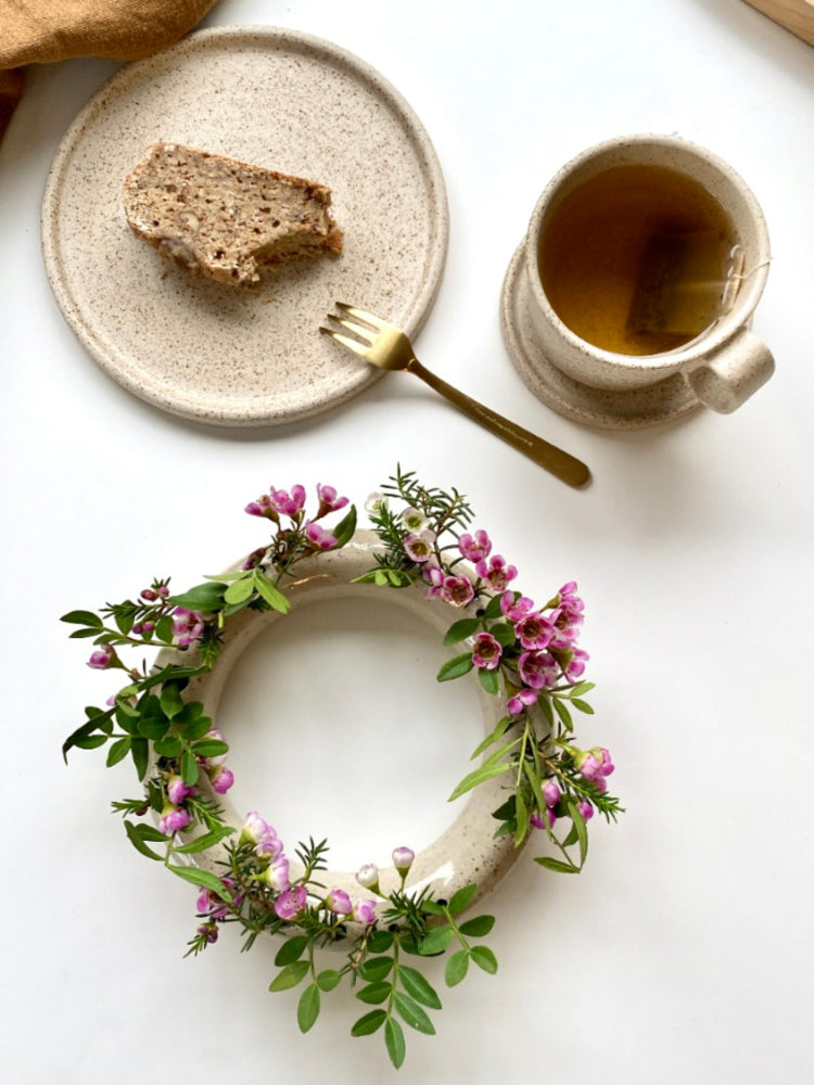 Beige ronde bloemenvaas met roze bloemen met een cakebord ernaast met cake en een mok met thee, bovenaanzicht
