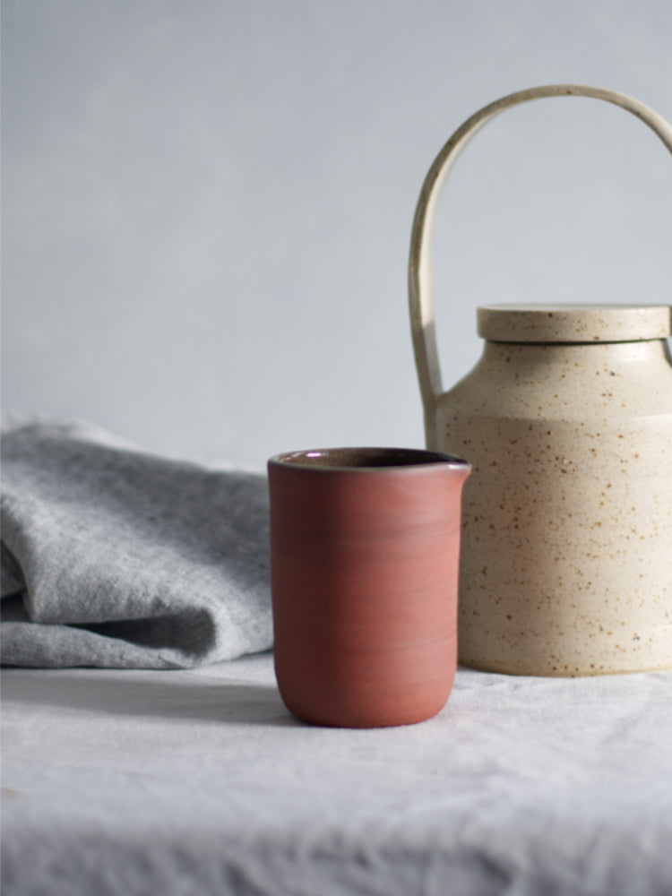 Rood schenkkannetje op een grijs tafelkleed, naast een theepot met een theedoek ernaast