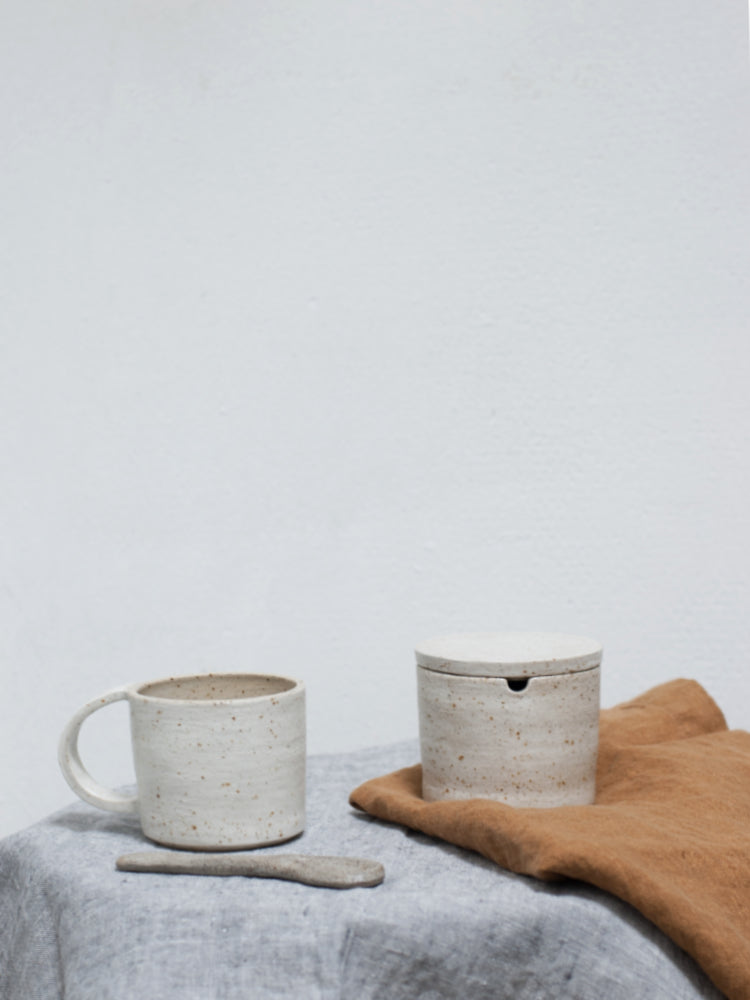 Handgemaakte suikerpot van keramiek met een handgemaakte mok met dezelfde afwerking ernaast op een tafelkleed en een theedoek ernaast.