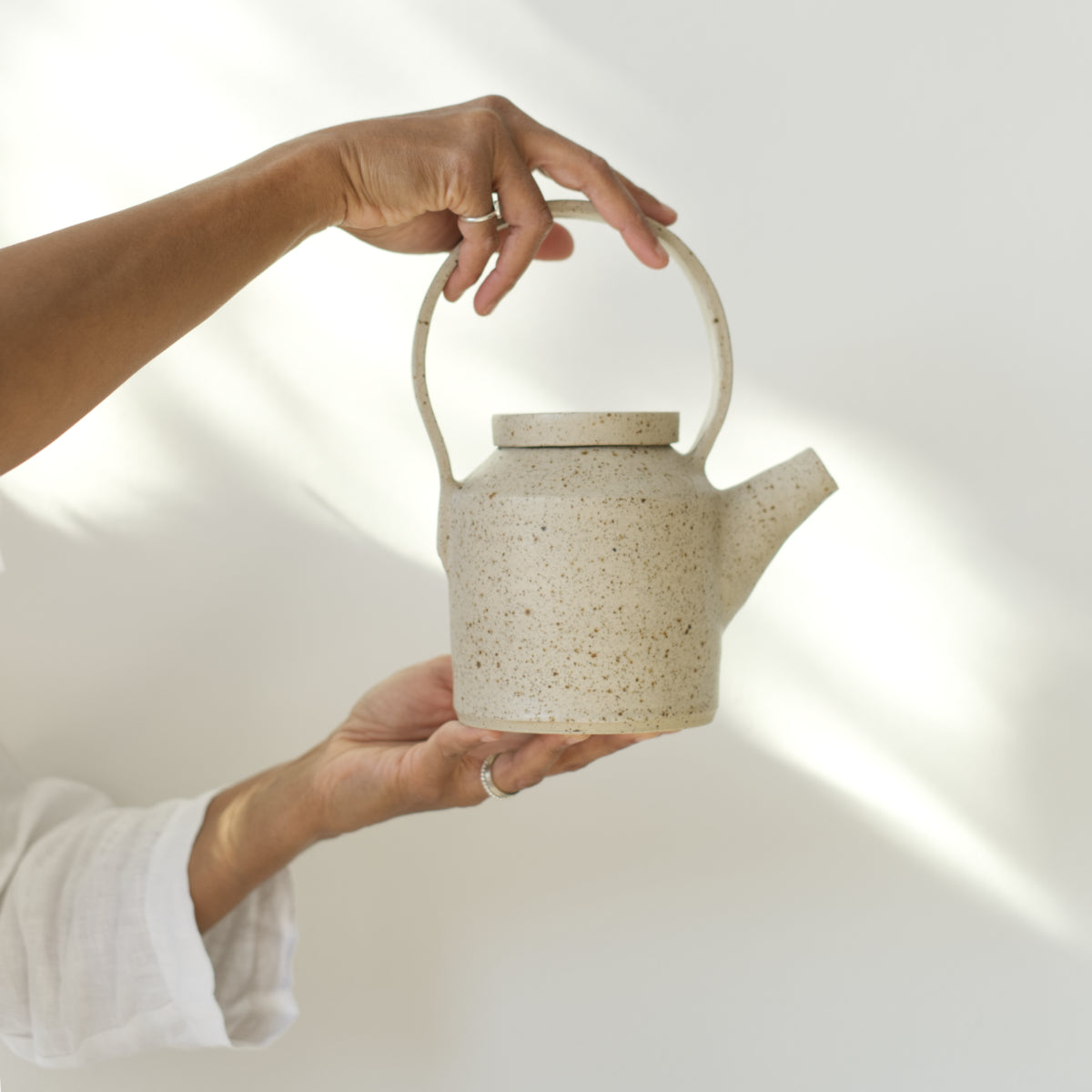 Handgemaakte gespikkelde keramische theepot van Imre Bergmann, vastgehouden in twee handen voor een witte muur met zachte schaduwen op de muur. 