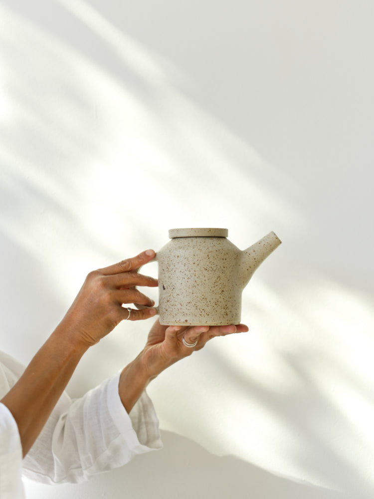 Twee handen die een handgemaakte theepot vasthouden voor een witte muur met daarop allemaal schaduwen van zon en bladeren