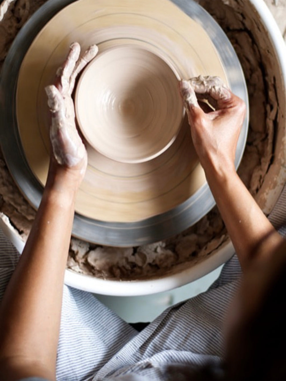 Bovenaanzicht van twee handen die werken achter een pottenbakkersdraaischijf met klei