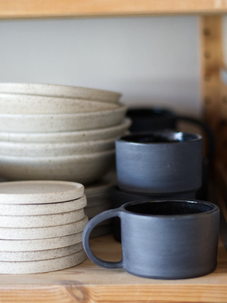 lage zwarte handgemaakte mok in op een plank in het atelier naast lichtgespikkelde kommen en schotels