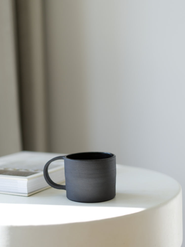 Handgemaakte zwarte keramieken mok in slow living stijl op een tafeltje met een boek erachter